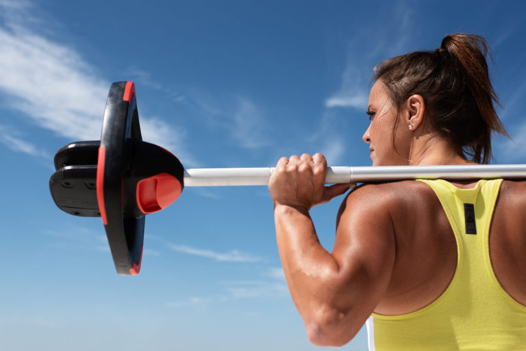 Kerri Anderson, a Les Mills National Presenter and fitness instructor, holds a barbell on her shoulders as she looks off into the sky. She is bathed in sunshine.