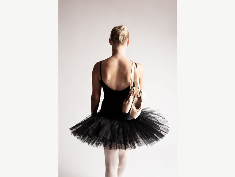 A ballerina stands facing away from the camera, holding her pointe shoes over her shoulder.