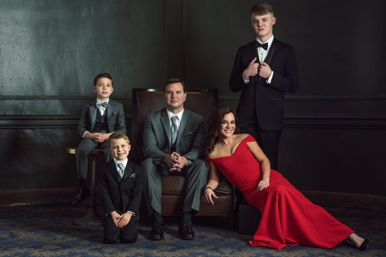 Family portrait with dramatic lighting, Dad is seated in a leather chair, surrounded by the boys and mother is seated near the ground, leaning on the chair in a red dress that pops.