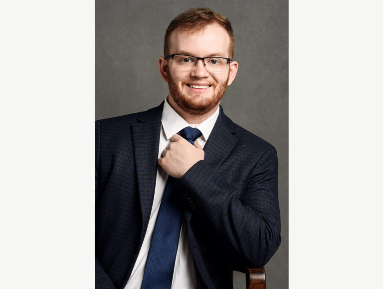 Personal branding head shot of a young professional man who adjusts his tie as he smiles with his audience. 
