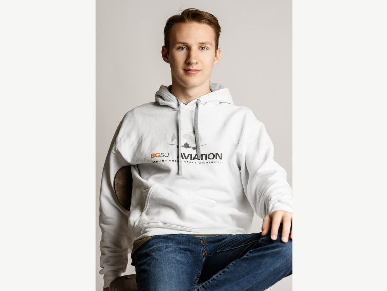 Editorial style photograph of a high school senior boy with understated confidence who relaxes in his chair as she shows off his college hoodie.