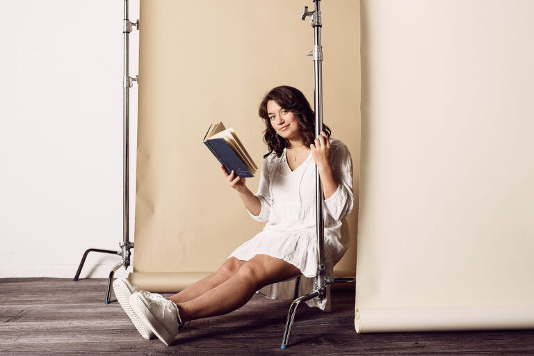 A high school senior girl poses with a book on a layered backdrop in studio during her photo session.