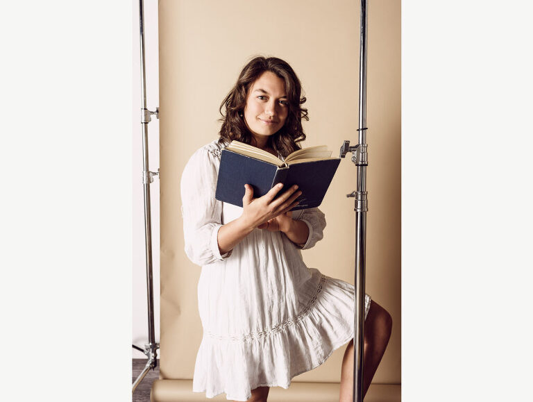 A high school senior girl poses with a book on a layered backdrop in studio during her photo session. One of her favorite books is The Alchemist, a novel about achieving one's destiny.
