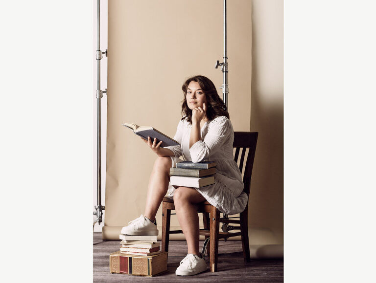 A studious high school senior girl poses with stacks of books on a layered backdrop in studio during her photo session.  One of her favorite books is The Alchemist, a novel about achieving one's destiny.