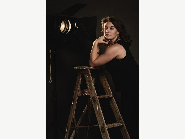 A high school senior girl poses with a ladder and an old fresnel light during her photo session in studio.