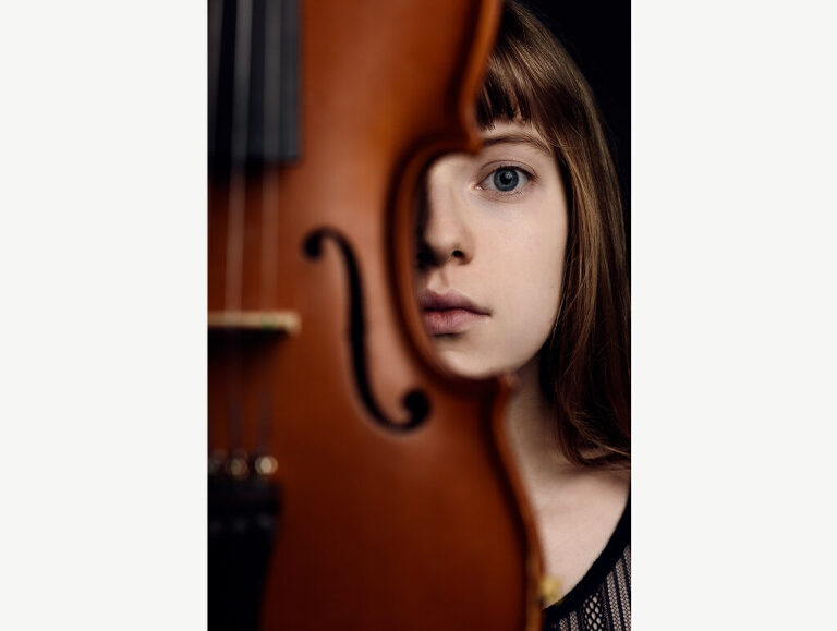 Studio photograph of a young violin player whose face is framed by her instrument. 