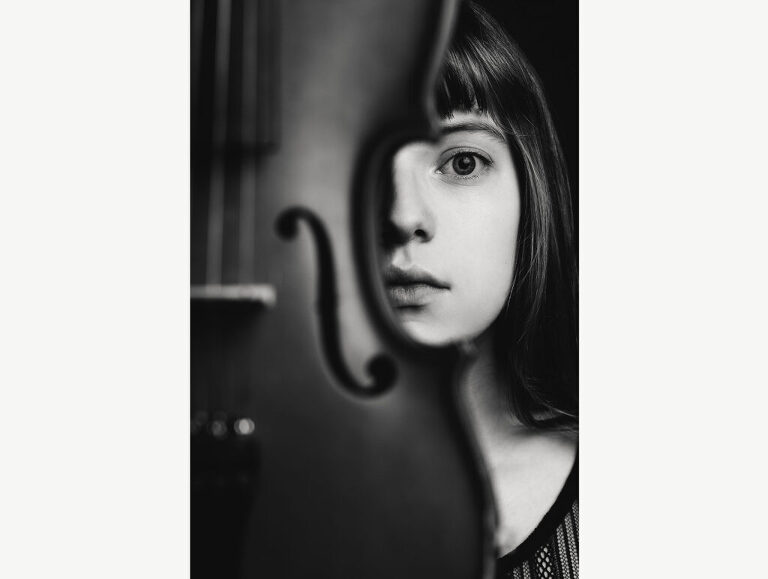 Black and white studio photograph of a young female violin player whose face is framed by her instrument. 