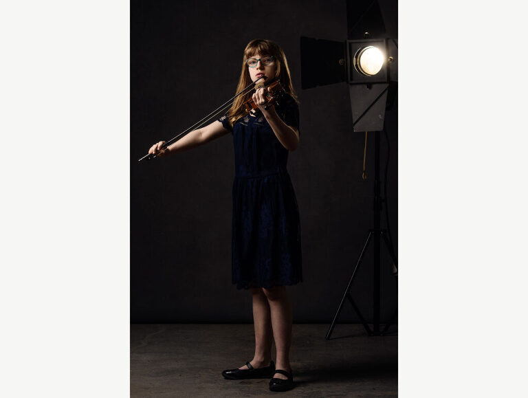 Studio photograph of a young female violin player standing in the spotlight as she plays.