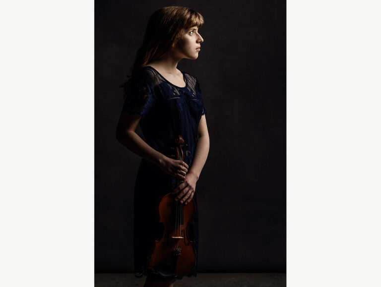 A young girl faces the light as she holds her violin at her side in a contemplative manner.