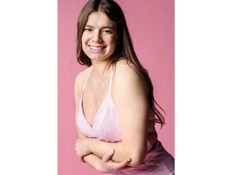 Portrait of a high school senior girl wearing her pink prom dress, set against a pink backdrop.