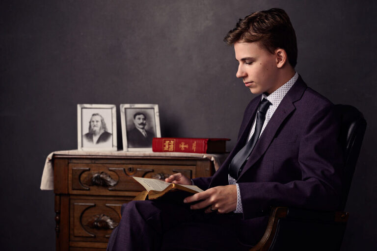 A studious high school senior boy reads a book in a study setting, with  a bible and framed portraits of his ancestors in the background